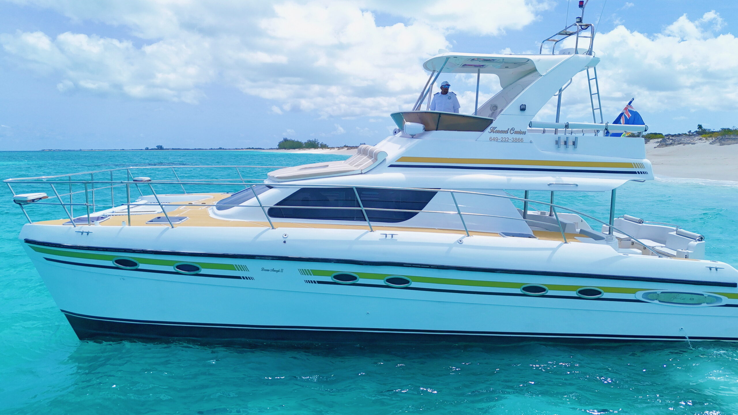 A sleek white catamaran with a yellow and green stripe is anchored in vibrant turquoise waters, with a crew member visible at the helm.