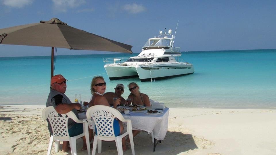 Group enjoying a beachfront meal under an umbrella, with a catamaran anchored in the turquoise water behind them. Relaxed, tropical atmosphere.