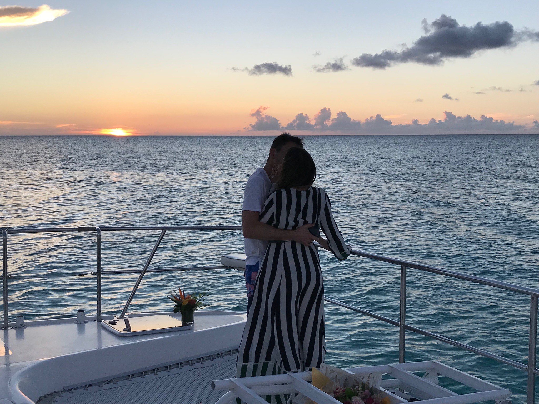 Couple embracing on a yacht at sunset, with calm ocean waters and colorful clouds, creating a romantic atmosphere.