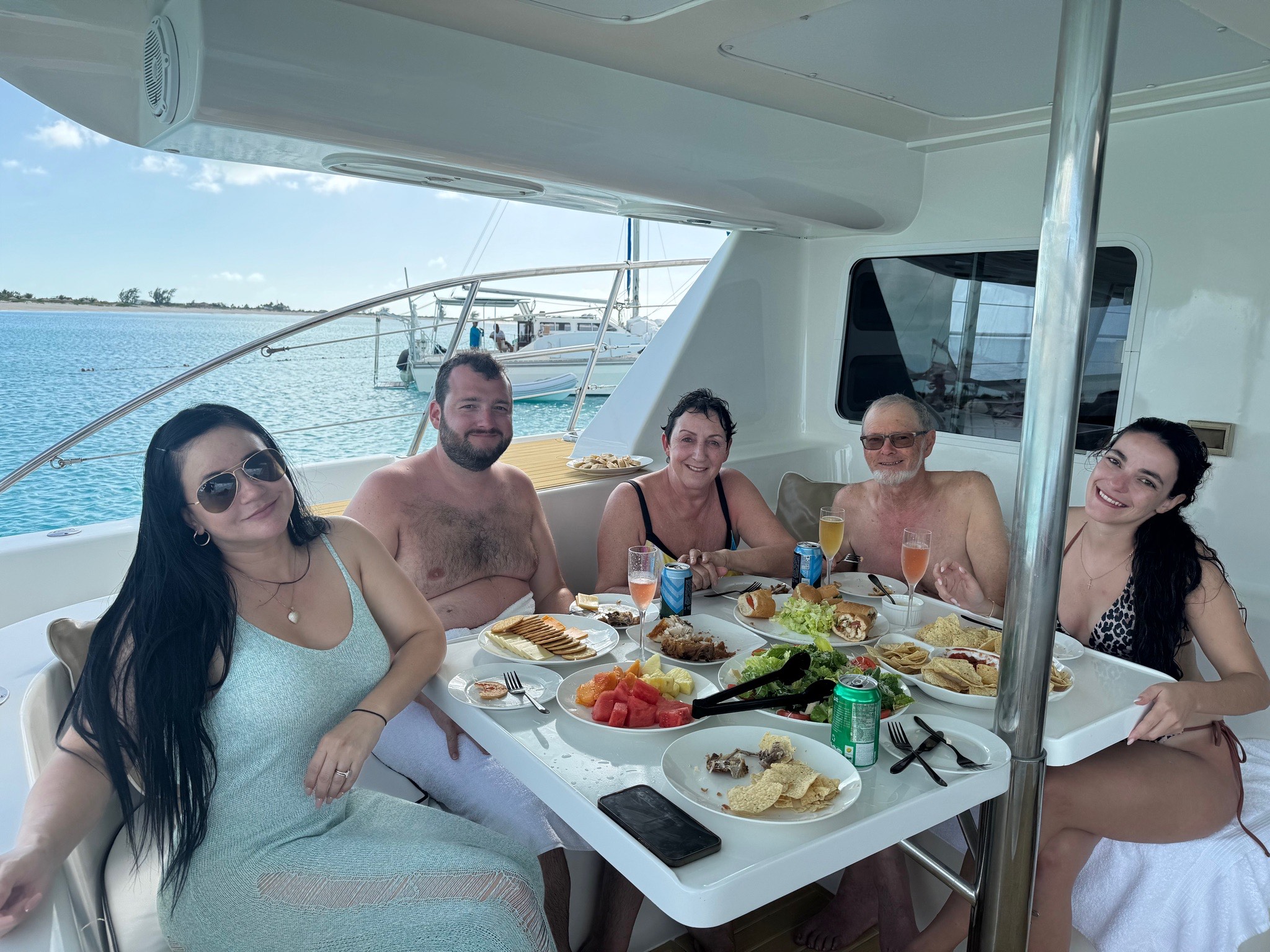 Five people enjoy a casual meal aboard a yacht, featuring a variety of dishes and drinks, with a scenic ocean view in the background.