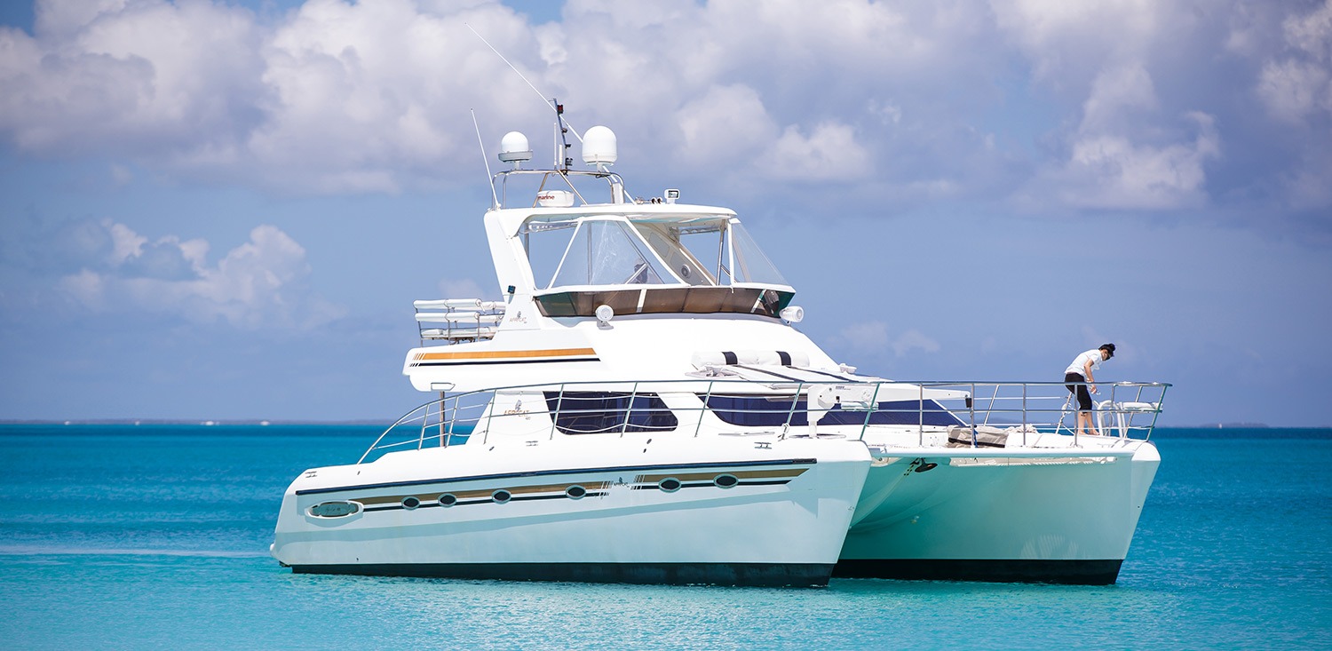 A sleek catamaran anchored in turquoise waters, with a person on board tidying up under a partly cloudy sky, emphasizing a relaxed maritime lifestyle.